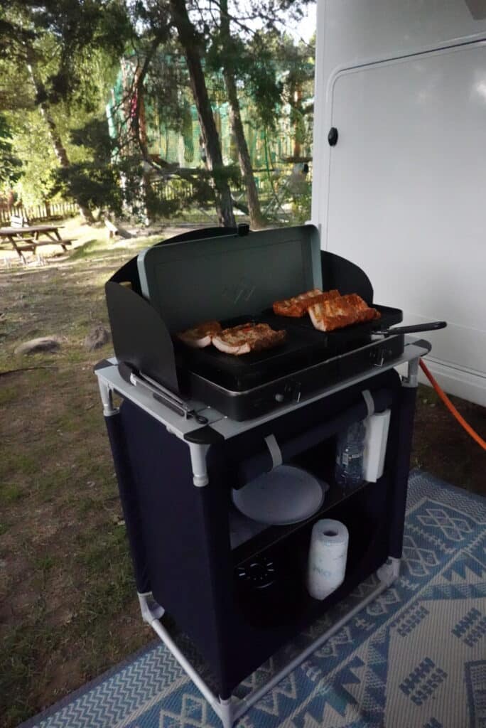 Barbecue met vlees op de grill, opgesteld naast een Reislust camper op een camping, met bomen en een picknicktafel in de achtergrond. Camper met buitenkeuken