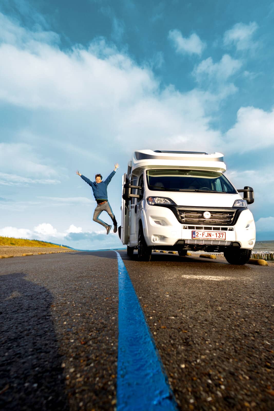 Een persoon springt naast een Reislust camper op een verlaten weg, met een heldere blauwe lucht op de achtergrond.