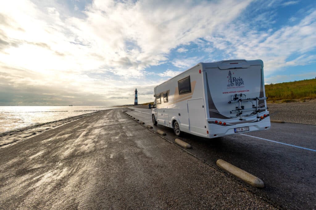 Achteraanzicht van een Reislust camper geparkeerd op een kade bij een vuurtoren, met een bewolkte lucht en een ondergaande zon.