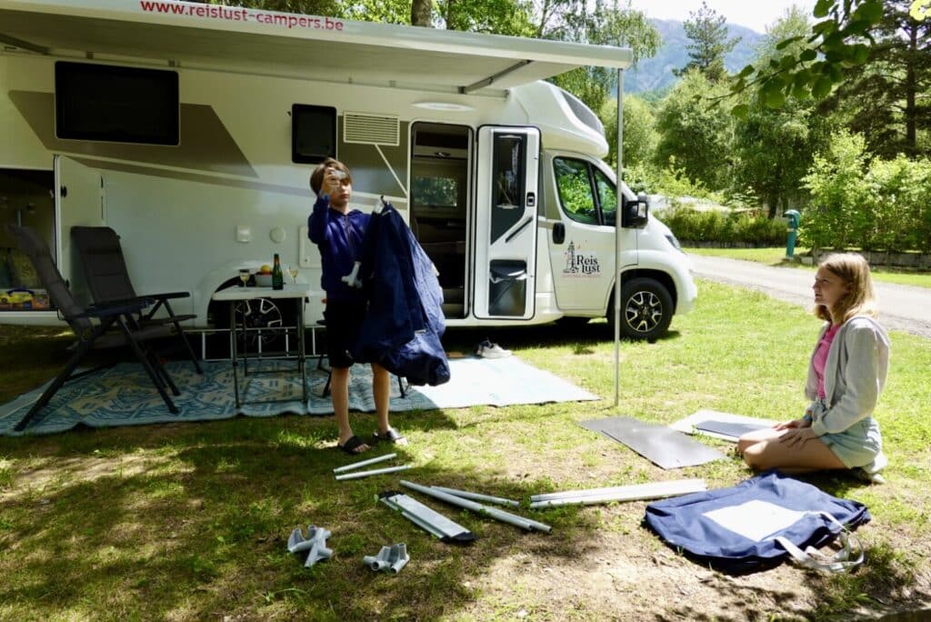 Twee personen aan het opzetten van een tent voor een Reislust camper op een camping, met stoelen en tafel onder de luifel.