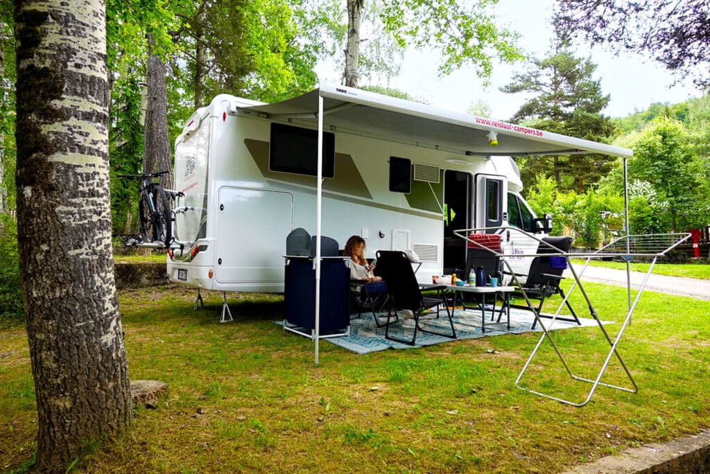 Reislust camper op een groene camping met een persoon zittend onder de luifel, omringd door kampeermeubelen. Camper met buitenkeuken